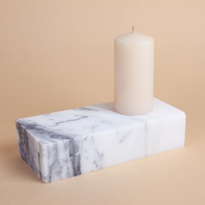 May include: A white pillar candle sits on a rectangular marble candle holder with grey veining.