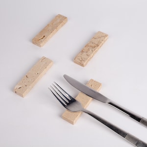 May include: Four rectangular, beige stone knife rests. A stainless steel fork and knife rest on one of the stone rests. The knife has the word "Camino" printed on the blade.