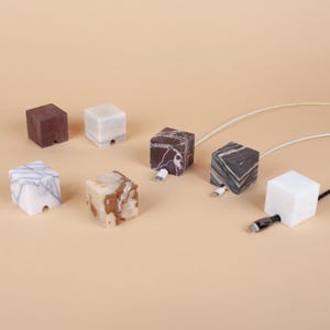 May include: Marble cube cable tidies in a range of colours and patterns, including white, brown, and grey. Each cube features a slot for a charging cable to pass through, with some cables connected.