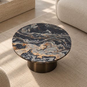 Op de afbeelding: Een ronde salontafel met een cirkelvormig blad van donkerblauw, zwart en goud geaderd marmer. De tafel wordt ondersteund door een massieve, cilindrische voet met een metallic bronzen afwerking, wat zorgt voor een stabiele basis.