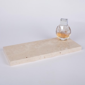 May include: A small, round perfume bottle with a silver cap sits on a rectangular, beige marble tray. The perfume bottle is clear with a faceted design, and the liquid inside is amber-colored. The tray has a smooth surface and a natural stone appearance.