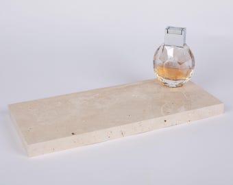 May include: A rectangular beige travertine stone tray with a porous, natural surface texture. Resting on the right side of the tray is a clear, spherical glass perfume bottle with a silver metallic cap containing amber liquid.