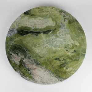 May include: A circular slab of polished green stone featuring a complex, marbled pattern with deep forest green, light olive, and creamy white veins. The surface displays natural mineral variations and smooth textures across the entire round form.