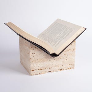 May include: A book stand made of beige travertine stone with a book resting on top. The book is open and has a black cover.