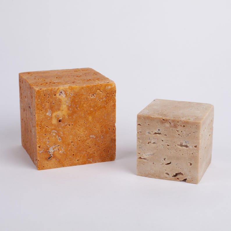 May include: Two square blocks of travertine stone. The larger block is a warm yellow color, while the smaller block is a light beige color.