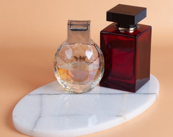 May include: Two perfume bottles on a white marble tray. The bottle on the left is clear glass with a faceted design and a silver cap. The bottle on the right is a dark red glass bottle with a black cap.