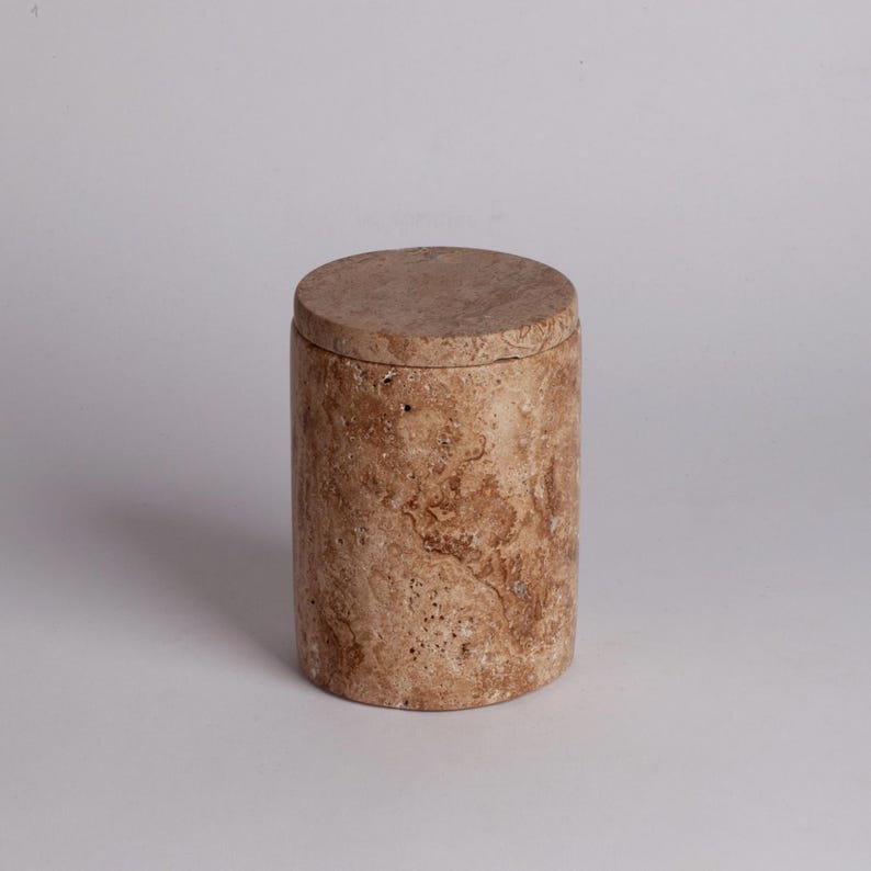 May include: A cylindrical, beige travertine stone container with a lid. The container has a natural, marbled pattern with brown and beige tones.