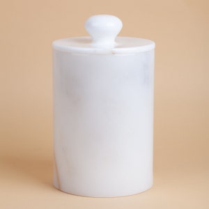 May include: White marble cylindrical container with a white knob lid. The container is smooth and has a subtle marbled pattern.