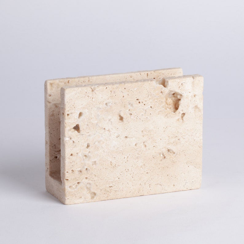 May include: A beige stone napkin holder with a rough, textured surface. The holder has a rectangular shape with an open top and a small opening on the side.