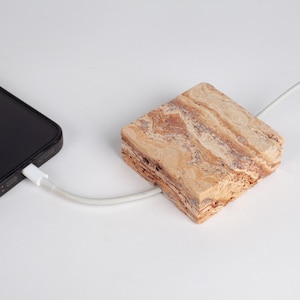 May include: A square-shaped wireless charging pad made of tan and brown marbled stone with a rough, porous side texture. A white charging cable connects the stone pad to a black smartphone, which is partially visible on the left side of the frame.
