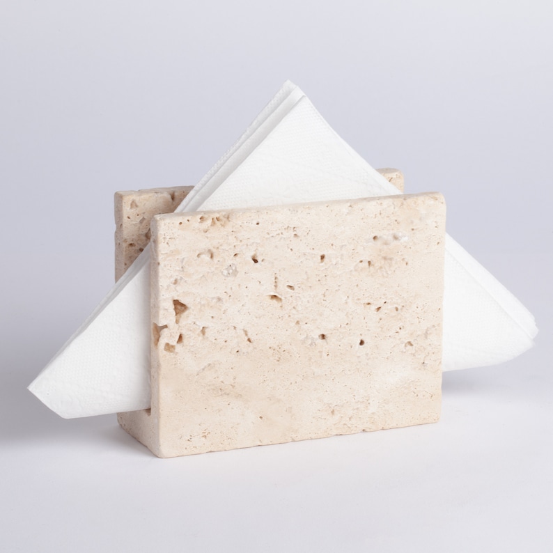 May include: A beige travertine napkin holder with a white napkin inside.