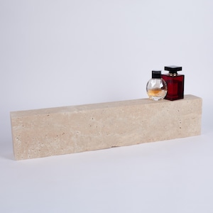 Può includere: Una mensola rettangolare in pietra beige con due flaconi di profumo. Uno è rotondo e trasparente con tappo nero, l'altro è un flacone quadrato rosso scuro con tappo nero. La mensola è in pietra chiara.