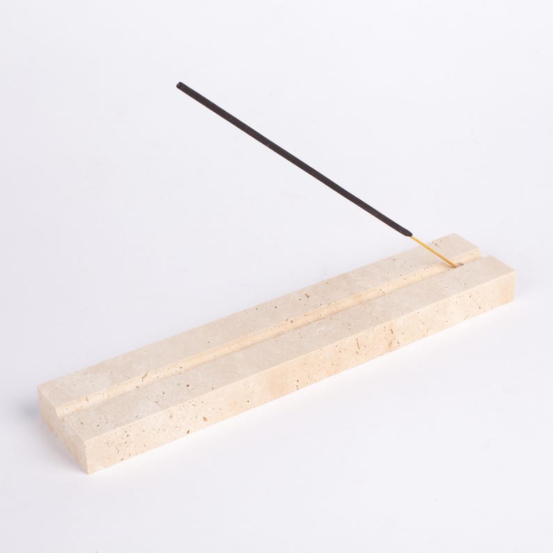 Pode incluir: Um porta-incenso retangular de pedra bege com uma ranhura para segurar varetas de incenso. Uma vareta de incenso preta com ponta dourada &eacute; inserida no suporte, angulada para cima. O suporte &eacute; feito de pedra de cor clara.