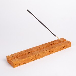Pode incluir: Um porta-incenso retangular, poroso, marrom-alaranjado com duas ranhuras paralelas. Um palito de incenso preto com ponta dourada &eacute; inserido em uma das ranhuras, inclinado para cima. O suporte &eacute; feito de um material texturizado.