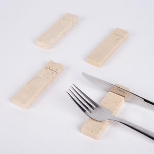 May include: A collection of beige rectangular stone knife rests with a fork and knife. The knife rests are made of a textured stone material. The knife has the word "Camino" printed on the blade.