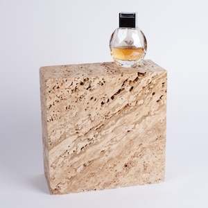May include: A clear glass bottle of perfume with a silver cap sits on a beige travertine stone block.