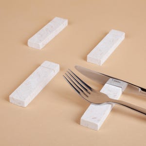 Puede incluir: Cuatro reposacuchillos rectangulares de mármol blanco con un patrón moteado. Un tenedor y un cuchillo descansan sobre uno de los reposos. El cuchillo tiene la palabra "Camino" impresa. El fondo es de color beige liso.