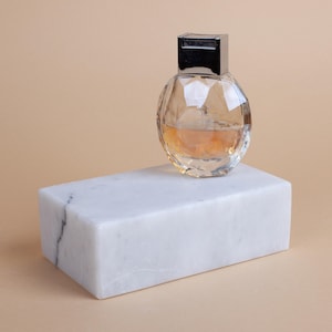 Könnte beinhalten: Ein klarer, facettierter Parfümflakon mit silberner Kappe, gefüllt mit bernsteinfarbener Flüssigkeit, ruht auf einem weißen Marmorblock. Der Hintergrund ist einfarbig hellbeige. Die Flasche hat ein geometrisches Design.