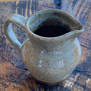 May include: A speckled brown and gray ceramic pitcher with a handle. The pitcher is filled with a dark liquid and is sitting on a wooden surface.