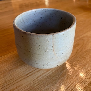 May include: A handmade, speckled ceramic cup with a cylindrical shape. The cup is a light beige color with dark brown speckles and a smooth, unglazed exterior. The cup is suitable for beverages.