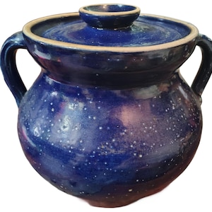Op de afbeelding: Een blauwe keramische pot met een deksel en twee handvatten. De pot heeft een gespikkeld patroon en een glanzende afwerking. Het deksel heeft een kleine knop bovenop. De pot is diepblauw met een lichtere blauwe rand.