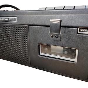 May include: A vintage black Sanyo portable cassette player with a handle. The player has a speaker grill, a cassette tape slot, and control buttons. The text "SANYO PORTABLE CASSETTE" is visible on the front.
