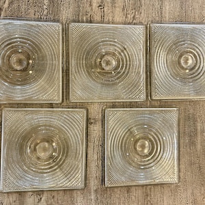 May include: Five clear, square glass tiles with a concentric circle design radiating from a central point. The tiles are arranged on a light-colored wooden surface, showcasing their transparent quality.