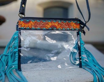 May include: Clear crossbody bag with turquoise fringe and a sunflower design on the top. The bag has a black strap and a clear front, perfect for concerts and events. The bag is made of leather and has a zipper closure.