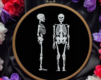 May include: A black and white embroidery hoop with two skeletons embroidered on a black fabric background. The skeletons are standing side by side, one is facing forward and the other is facing to the side. The hoop is surrounded by a variety of colorful flowers.