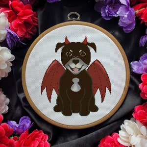 Könnte beinhalten: Ein brauner und schwarzer Hund mit roten Teufel Flügeln und Hörnern ist auf einem weißen Stoff in einem hölzernen Stickrahmen gestickt. Der Rahmen ist von lila, weißen und roten Blumen umgeben.