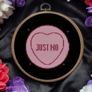 May include: A pink heart-shaped candy with the text "JUST NO" in black letters, framed in a wooden hoop against a black background.