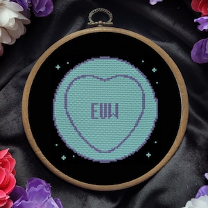 May include: A cross-stitch embroidery hoop with a black background and a light blue candy heart with the word "EWW" in purple lettering. The candy heart is surrounded by small white stars.