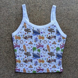 May include: White tank top with a colorful all-over print featuring cartoon characters, cruise ships, anchors, surfboards, and tropical elements. The top has a white trim and straps. The word "CAT" is visible on the print.