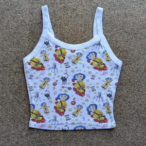 May include: White vest top with a repeating pattern featuring a cartoon character in a yellow dress and blue hat, along with black cats, stars, and other whimsical elements. The vest top has a scoop neck and thin straps.