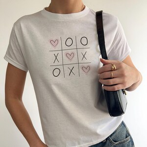May include: White t-shirt with a graphic of a tic-tac-toe board with hearts and X's.