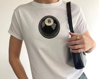 May include: White t-shirt featuring a black and white illustration of a pool ball with the number 8 on it.