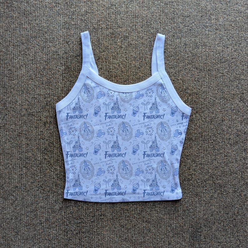 May include: A light blue tank top with a white trim and straps. The tank top features a repeating pattern of castles, Mickey Mouse, fireworks, and the word "Fantasmic!".