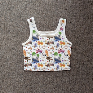May include: White tank top with a colorful all-over print featuring cartoon characters, boats, anchors, palm trees, and surfboards. The top has a scoop neck and wide straps.