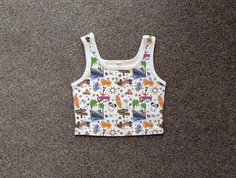 May include: White tank top with a colorful all-over print featuring cartoon characters, boats, anchors, palm trees, and surfboards. The top has a scoop neck and wide straps.