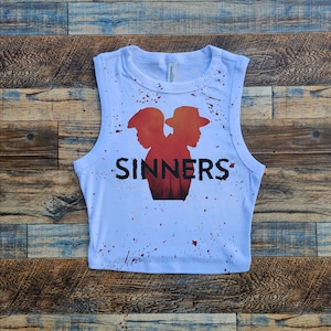 Puede incluir: Un top corto blanco sin mangas con un estampado gráfico de dos siluetas en tonos rojos y naranjas, con la palabra SINNERS impresa en negro debajo. La camiseta está decorada con salpicaduras de pintura roja en el frente y el cuello.