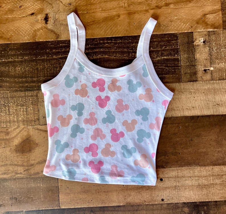 May include: White crop top with a pattern of pink, peach, and blue Mickey Mouse silhouettes. The tank top has a scoop neck and thin straps. The top is made of a soft, lightweight material.