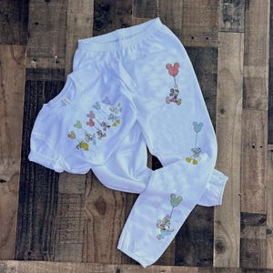 Pode incluir: Conjunto de camiseta e cal&ccedil;as de moletom brancas com personagens de desenhos animados segurando bal&otilde;es. A camiseta tem gola redonda e mangas curtas. As cal&ccedil;as de moletom t&ecirc;m c&oacute;s e punhos el&aacute;sticos. Os personagens incluem Mickey, Donald e Pateta.