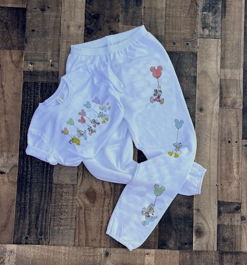 Pode incluir: Conjunto de camiseta e cal&ccedil;as de moletom brancas com personagens de desenhos animados segurando bal&otilde;es. A camiseta tem gola redonda e mangas curtas. As cal&ccedil;as de moletom t&ecirc;m c&oacute;s e punhos el&aacute;sticos. Os personagens incluem Mickey, Donald e Pateta.
