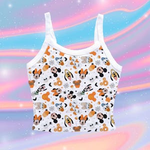 Puede incluir: Una camiseta de tirantes blanca con un estampado integral de personajes de Disney como Mickey Mouse, Minnie Mouse, Donald Duck y Pluto, intercalados con iconos de Halloween como maíz dulce, telarañas, murciélagos y calabazas naranjas.