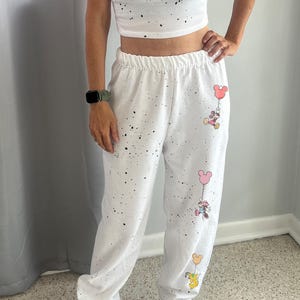 Pode incluir: Cal&ccedil;as de moletom brancas com respingos de tinta preta e personagens de desenho animado do Mickey Mouse segurando bal&otilde;es. As cal&ccedil;as de moletom t&ecirc;m c&oacute;s el&aacute;stico e fazem parte de um conjunto combinando com um top cropped.
