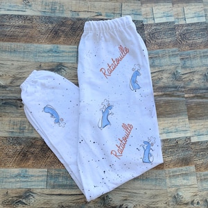 May include: White pajama pants with a repeating pattern of blue cartoon rats wearing chef hats and the word "Ratatouille" in red script. The pants have an elastic waistband and are speckled with black dots.