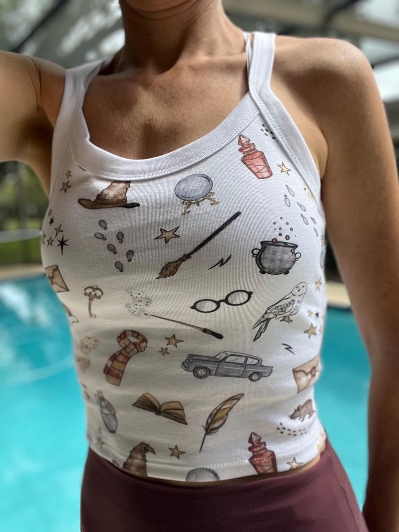 May include: White tank top with a print of various Harry Potter themed illustrations, including a magic wand, a potion bottle, a flying car, and a golden snitch.