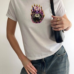 May include: White t-shirt with a black and white 8-ball graphic on fire. The 8-ball is surrounded by flames in shades of yellow, orange, pink, and purple.