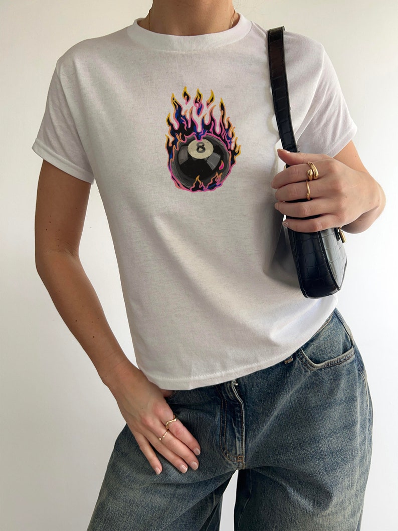 May include: White t-shirt with a black and white 8-ball graphic on fire. The 8-ball is surrounded by flames in shades of yellow, orange, pink, and purple.