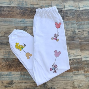 Pode incluir: Cal&ccedil;as de moletom brancas com personagens de desenhos animados segurando bal&otilde;es. As cal&ccedil;as t&ecirc;m c&oacute;s e punhos el&aacute;sticos. Os personagens de desenhos animados incluem Mickey Mouse, Minnie Mouse e Pluto. Os bal&otilde;es s&atilde;o rosa e bege.